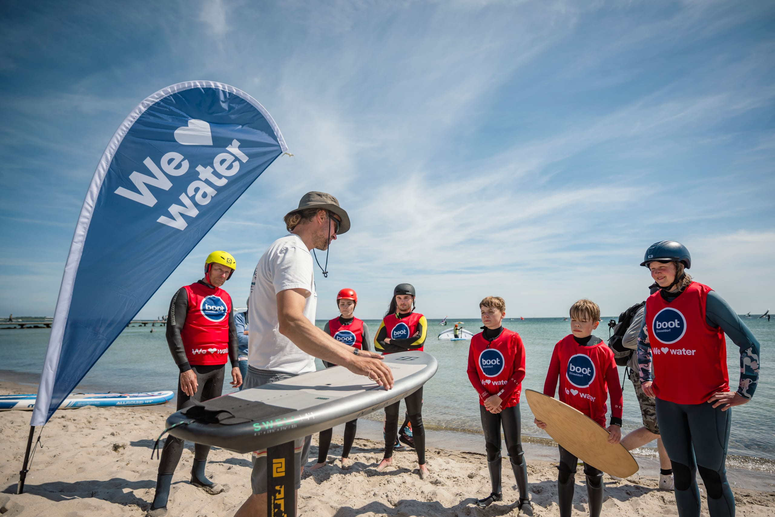 Surflehrer erklärt einer Gruppe in roten „boot Düsseldorf“-Leibchen am Strand ein Foilboard, daneben eine „We love water“-Fahne