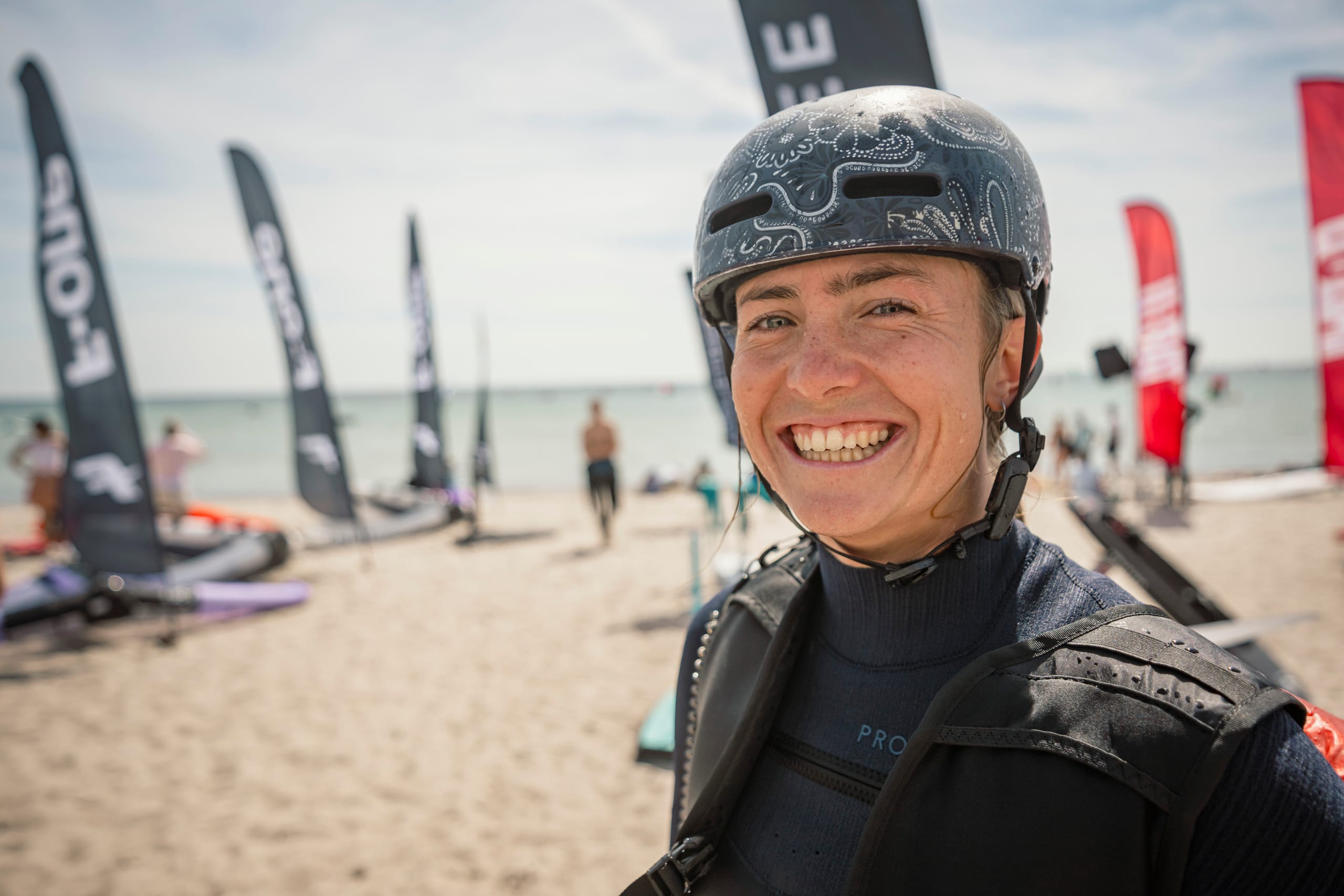 Lachende Surferin mit Helm und Neoprenanzug am Strand, im Hintergrund Ausstellerfahnen